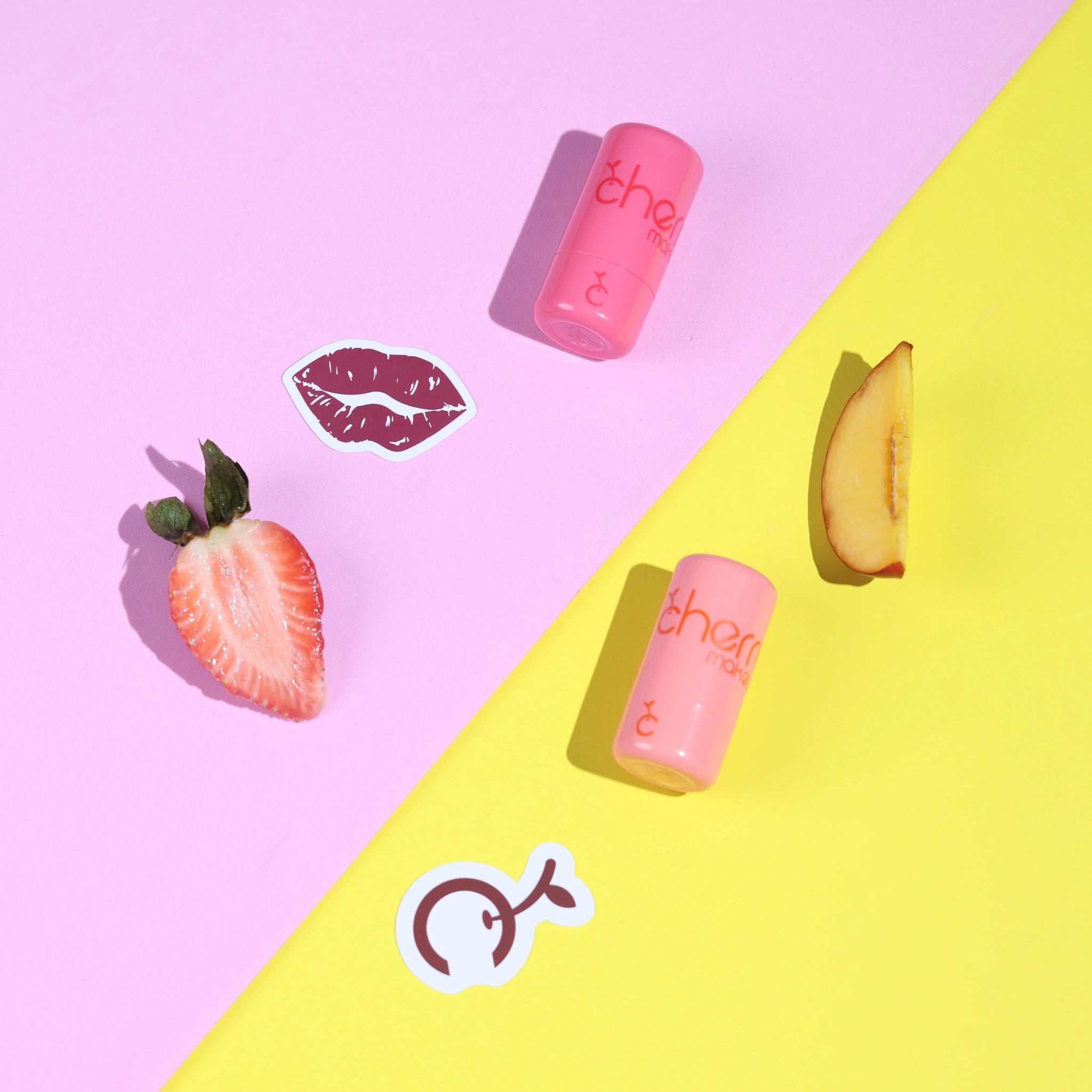 Two pink lip balms on a pink and yellow background with a strawberry and peach slice.